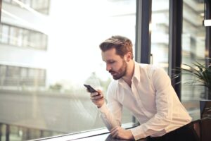 photo of man in white dress shirt holding phone near window